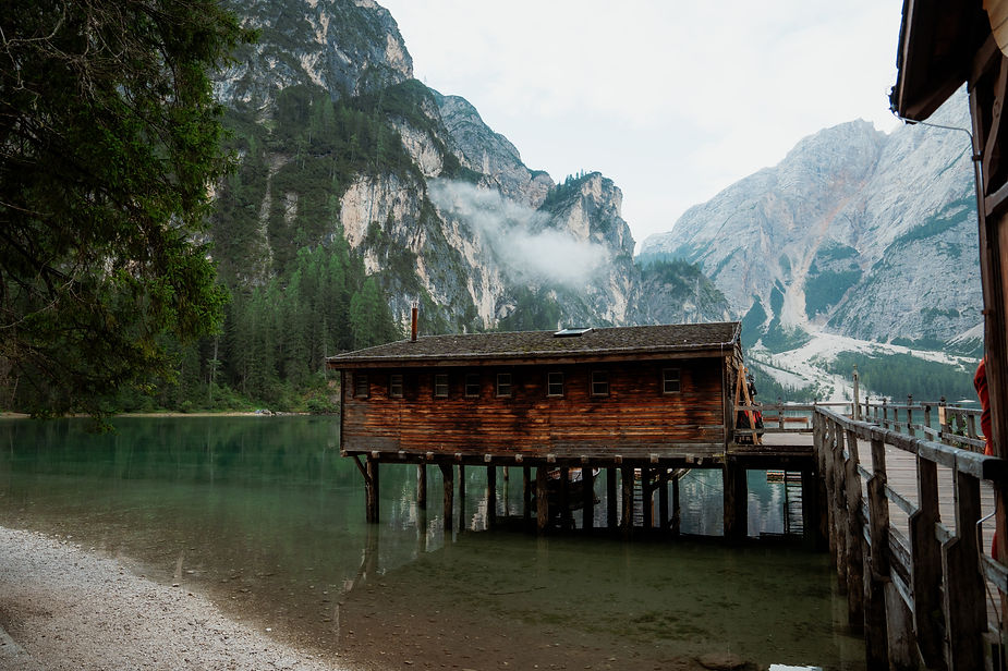 Wooden cabin on stilts over a calm lake, surrounded by towering mountains and trees. Misty clouds drift above, creating a serene atmosphere for a dolomites elopement at Lago di Braies