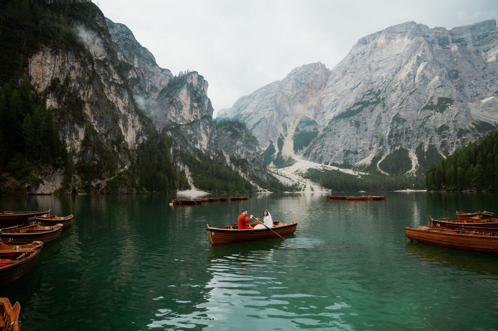 Two people row a wooden boat on a serene teal lake, surrounded by towering mountains and mist. Other empty boats float nearby during dolomites elopement