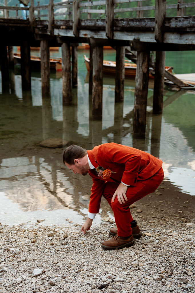 groom in red suit crouches by Lago di Braies, picking up a pebble. Wooden dock and boats in the background.
