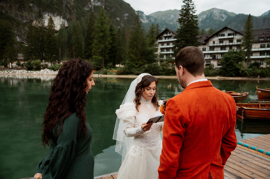during their dolomites elopement, bride reads vows to groom in orange jacket on the dock at Lago di Braies