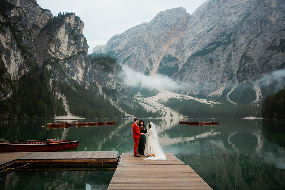 Bride and groom stand on a wooden pier by Lago di Braies, surrounded by misty mountains. The bride in white, groom in red. Peaceful atmosphere in the dolomites
