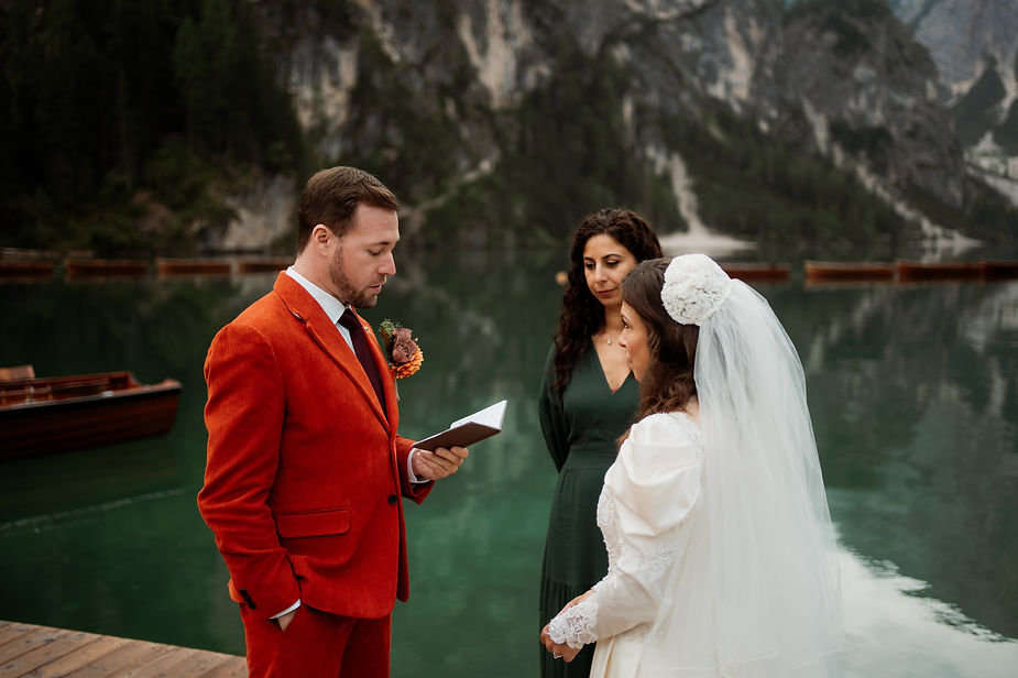 groom in orange suit reads vows to bride by Lago di Braies; officiant watches. Emotion: solemn during dolomites elopement