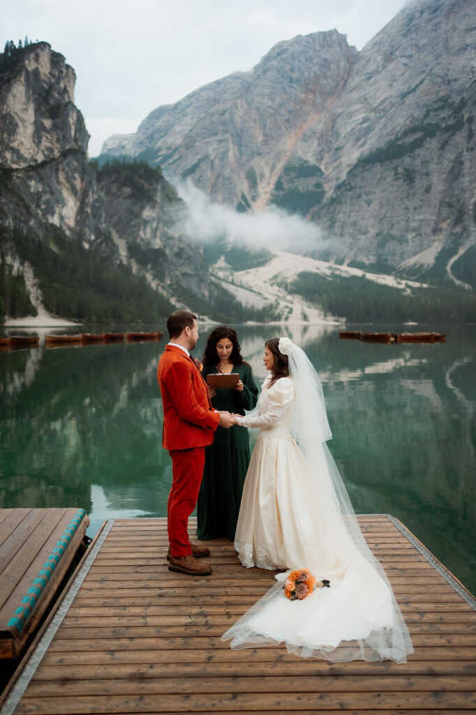 Bride and groom in wedding attire hold hands on a dock by Lago di Braies, with dolomites wedding officiant nearby and misty mountains in the background.