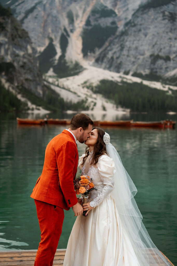 Bride and groom kiss by a Lago di Braies with italian alps. The groom wears a bright orange suit; the bride, a white dress with veil.