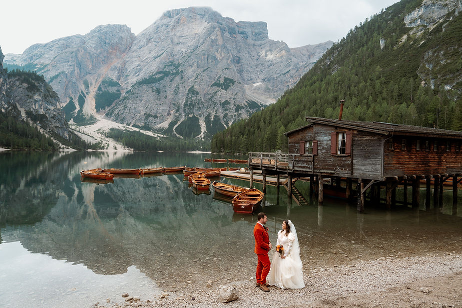 Bride and groom stand on the shore of Lago di Braies with boats and a wooden cabin nearby. Majestic italian alps in the background; serene and picturesque scene.