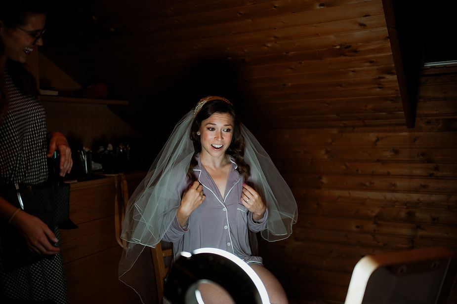 Bride in veil smiles, adjusting robe in cozy wooden room, lit by a ring light. Another person watches with a cheerful expression.