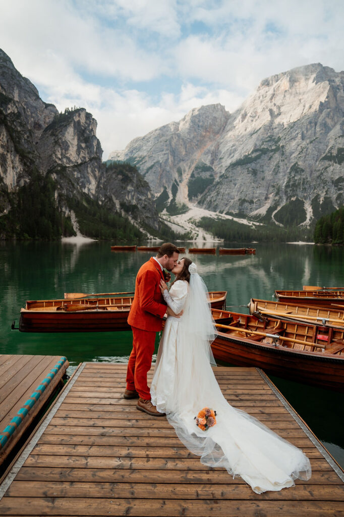 Bride and groom kissing on a Lago di Braies dock, surrounded by wooden boats and dolomites mountains.