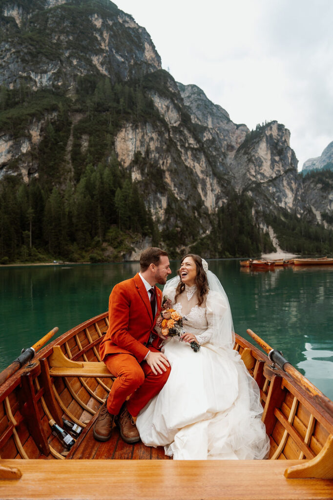 Bride and groom sit joyfully in a wooden boat on Lago di Braies, with towering dolomites mountains in the background of the elopement.