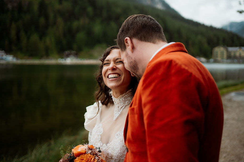 bride and groom laughing outdoors near a lake, with the woman in a white dress holding orange flowers and the man in an orange jacket during dolomites elopement at Lago di Braies