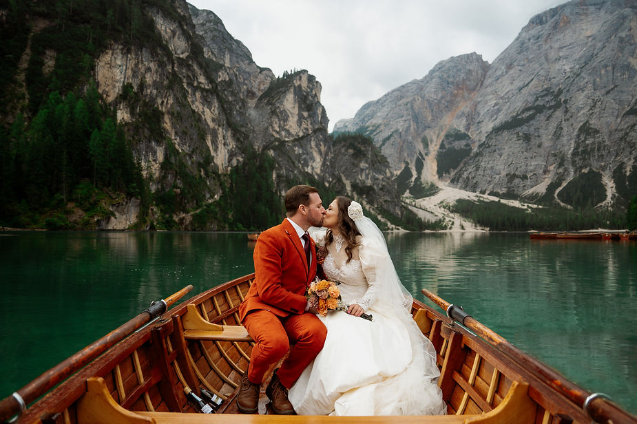 bride and groom in a wooden boat on the Lago di Braies, surrounded by mountains. The groom wears an orange suit; the bride, a white gown. Romantic scene for a dolomites elopement