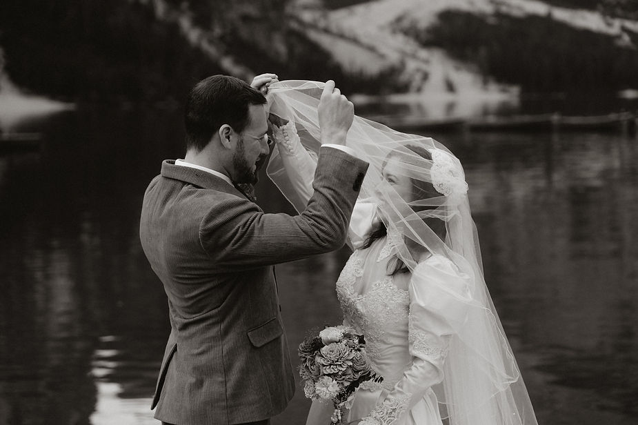 Groom lifts bride's veil by a serene lake in the italian alps. Bride holds a bouquet, wearing a lace dress. The mood is joyful and intimate.