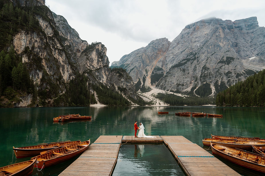Bride and groom stand on a dock by Lago di Braies, surrounded by wooden boats and majestic dolomites mountains. The mood is romantic and tranquil.