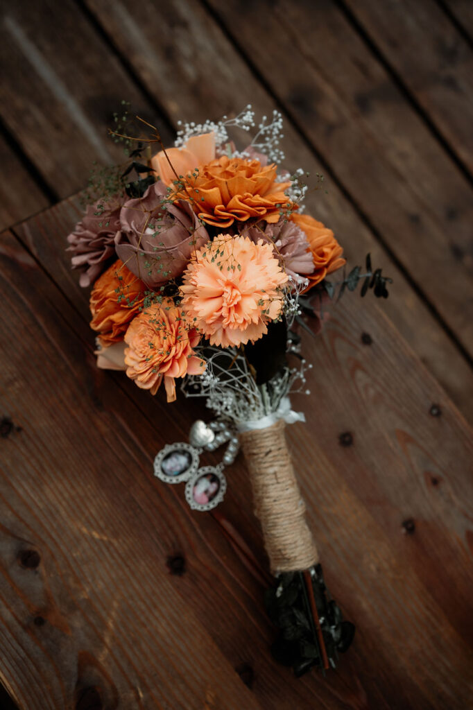 A wedding bouquet of orange and mauve flowers wrapped in twine with attached photo charms, on a wooden table. Rustic and elegant.