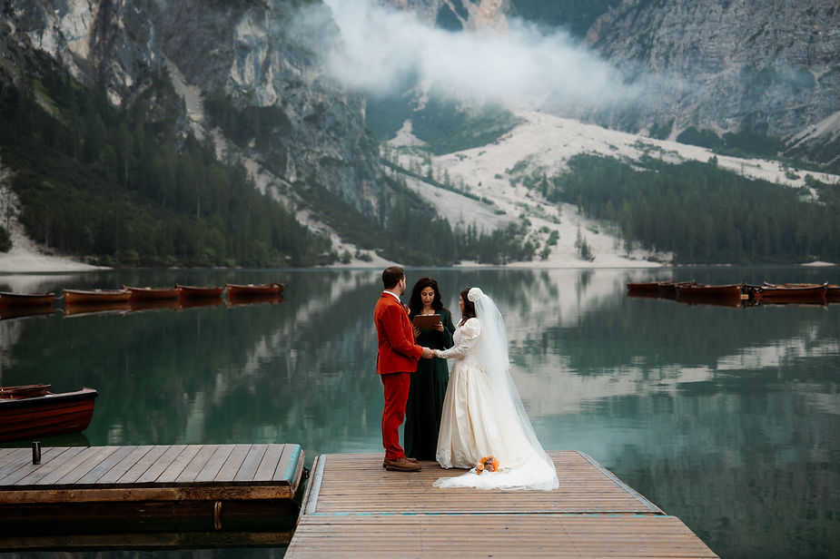 Bride and groom stand on a pier, holding hands, with an officiant by a misty mountain lake during wedding