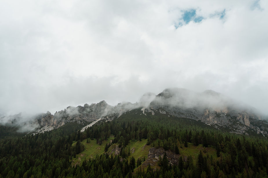 Foggy dolomites in italy with pine forest below, rocky peaks above, and clouds partially covering the summits under a cloudy sky.