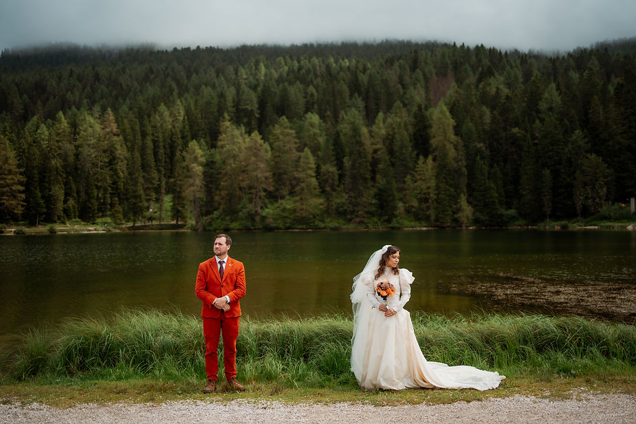 Bride in white gown holds bouquet, groom in orange suit stands beside her by Lago di Braies in italy. The sky is overcast.