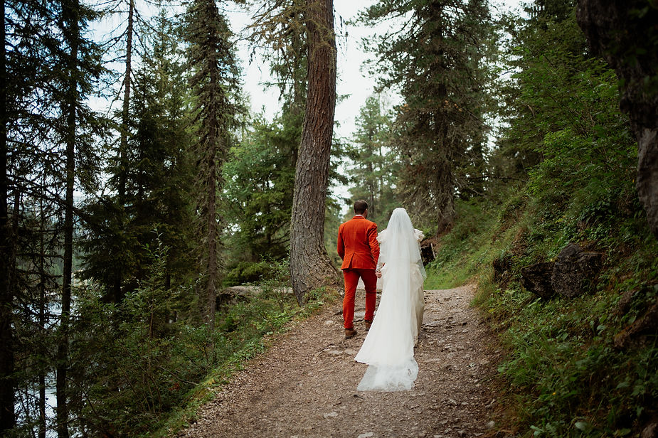Couple in wedding attire walks hand in hand on a forested path. Groom in red suit, bride in white dress and veil. Lush greenery surrounds them in the italian dolomites.