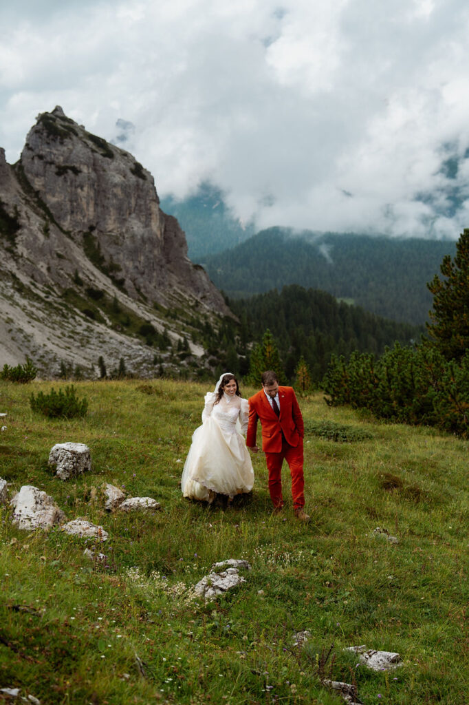 Bride in white dress and groom in red suit walk on grassy mountain slope, with italian dolomites in background, expressing joy and love during wedding photos