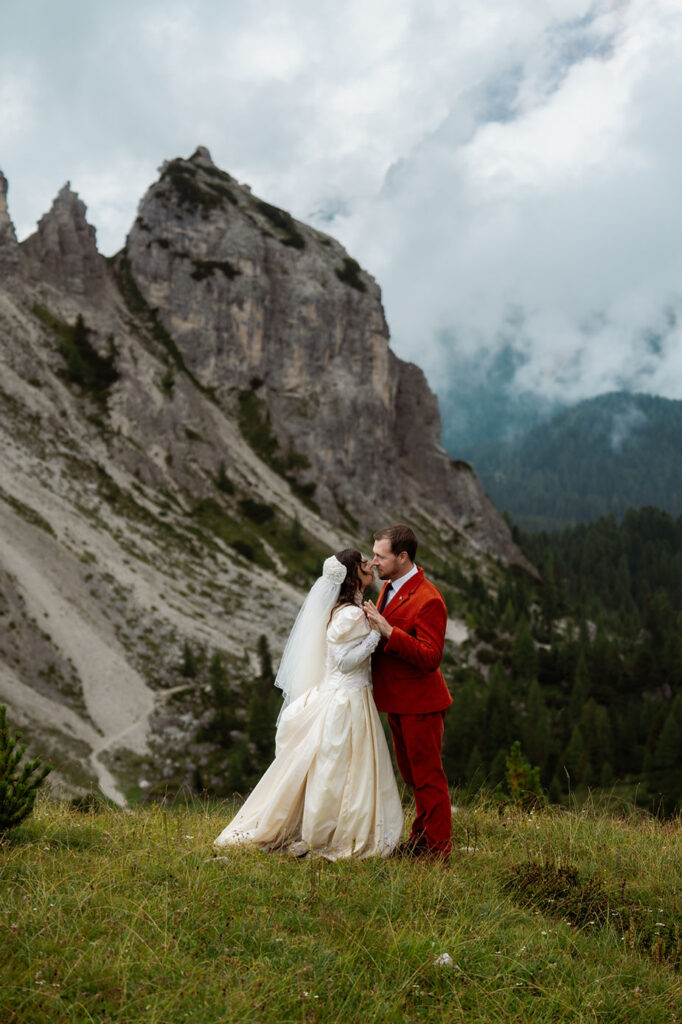 Bride and groom in red suit embrace on grassy hill with dolomites and cloudy sky in background. Romantic setting.