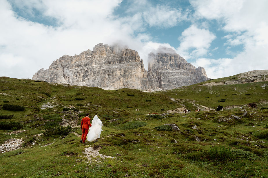 Bride in white, groom in red walk on grassy path, majestic mountains in background. Cows graze nearby under cloudy blue sky during dolomites wedding in italy. Peaceful mood.