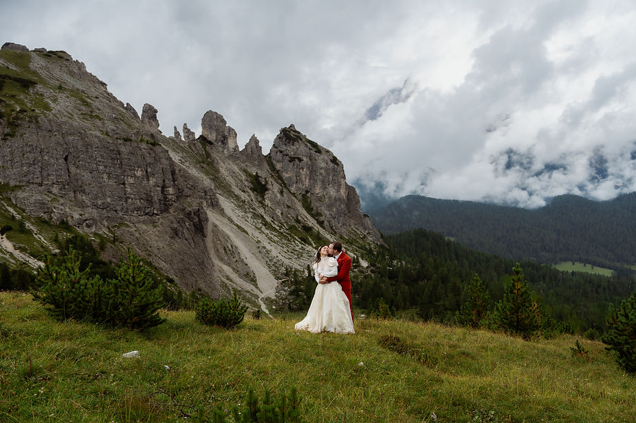 Bride and groom in red kiss on a grassy hill during dolomites elopement, surrounded by rocky cliffs and misty forest under cloudy skies. Romantic mood.