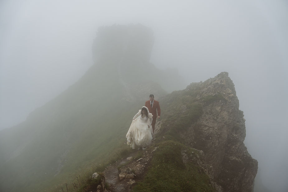 Bride in white dress and groom in red suit walk on a foggy trail in the dolomites. The misty backdrop creates a dreamy, mysterious mood.