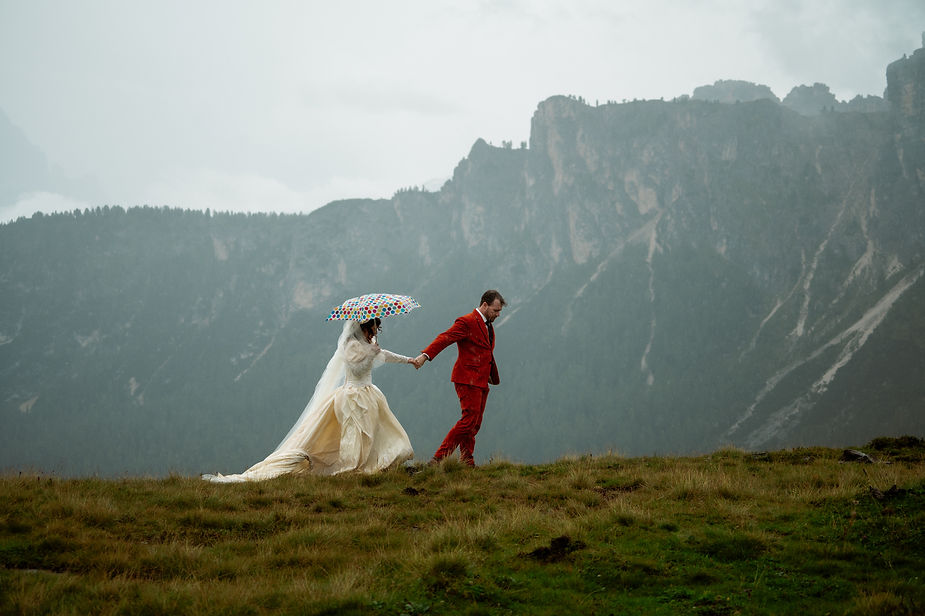A bride and groom hold hands on a grassy hill during dolomites wedding photos. The bride holds a polka dot umbrella. Misty mountains fill the cloudy background.