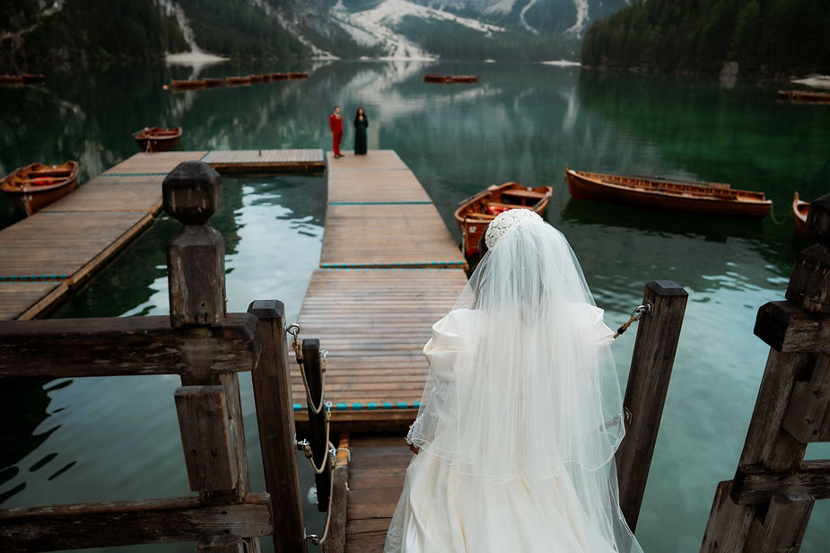Bride in white dress on wooden dock, facing a groom in red by Lago di Braies, surrounded by boats and lush mountains.