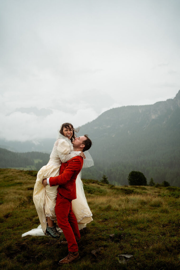 bride is joyfully lifted by groom on a grassy hill in the dolomites with misty dolomites mountains in the background.