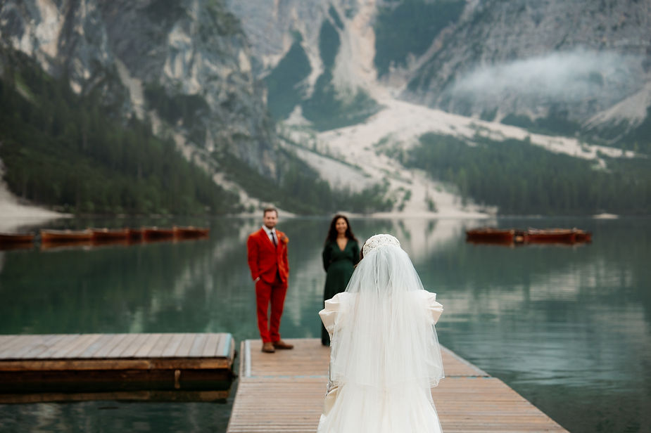 Bride in white dress faces couple on dock by a lake; groom in red suit. Misty mountain and boats in Lago di Braies during dolomites wedding