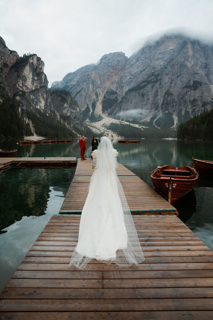 Bride in white gown walks on a dock towards two people by Lago di Braies with wooden boats during dolomites elopement in italy