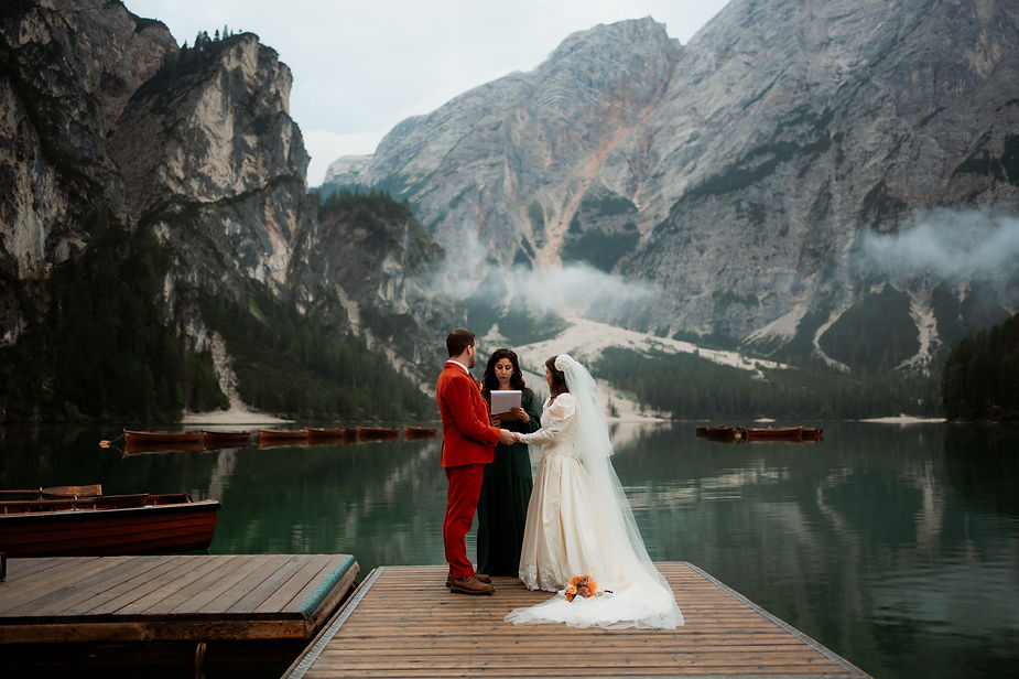 bride and groom holding hands on a dock by a lake, officiant reading. Misty mountains and boats on the Lago di Braies in italy