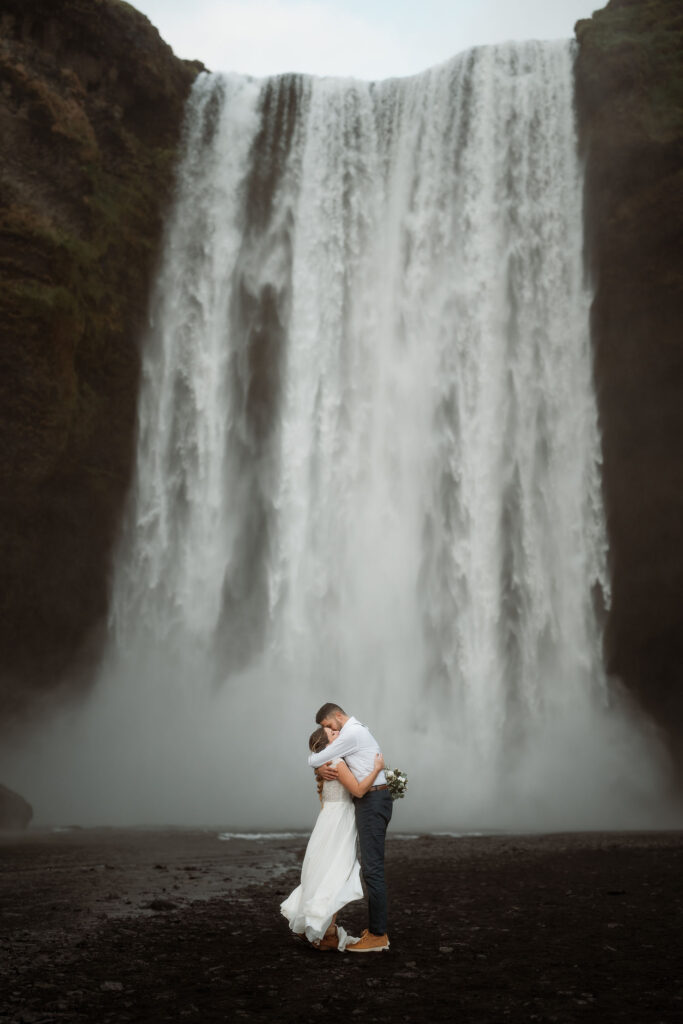 iconic iceland wedding location: A couple embraces in front of a large waterfall in iceland. The woman holds a bouquet and wears a white dress.