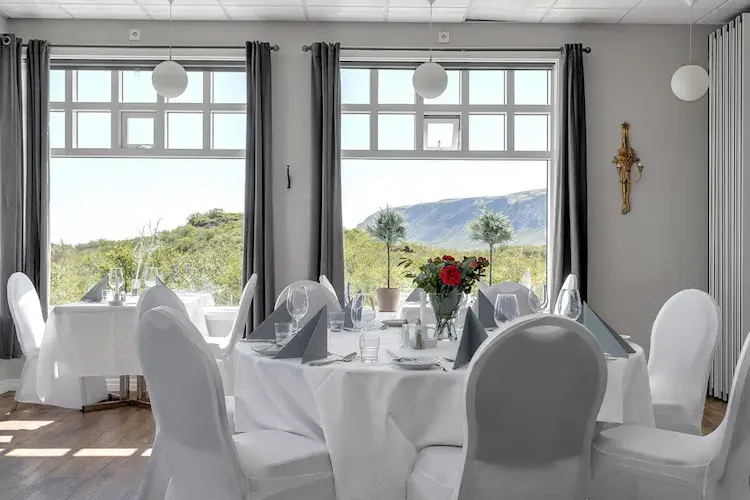 Elegant wedding venue with white-covered chairs and a round table set with glassware and flowers. Large windows show a scenic mountain view.
