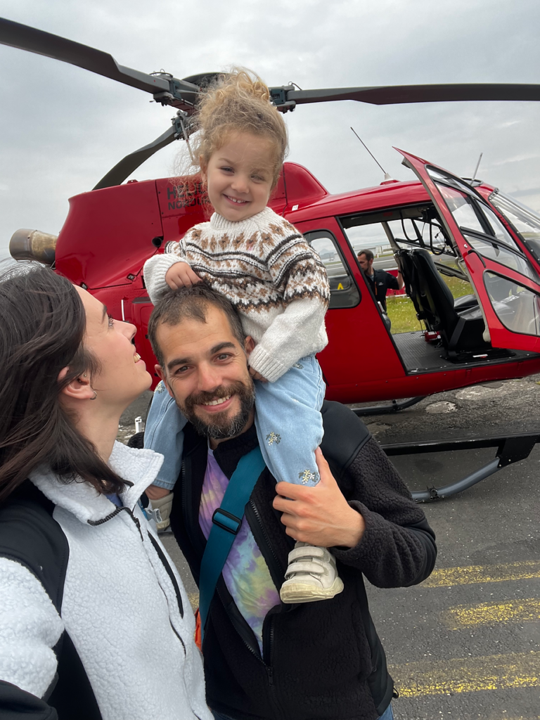 Smiling woman, man, and child in cozy sweaters by a red helicopter on a cloudy iceland day. Joyful mood, open helicopter door visible.