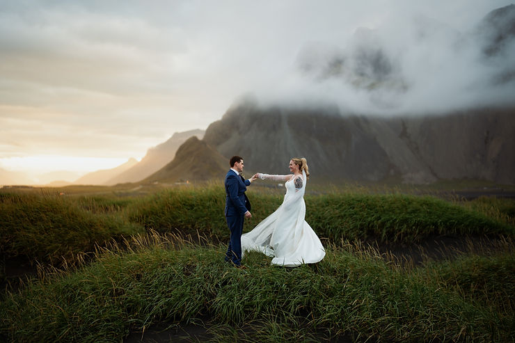 iconic iceland wedding locations: Bride in white dress and groom in blue suit dance on grassy hill at sunset, with misty mountains in the background.