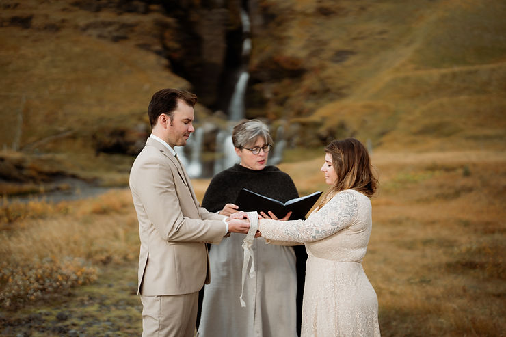 iconic iceland wedding locations: A bride and groom in beige attire holds hands for a ceremony led by a woman in black, set against a backdrop of a waterfall and grassy hills.