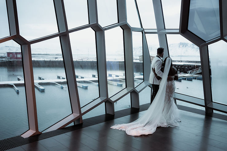 iconic iceland wedding locations: Bride and groom in elegant attire embrace by large geometric windows, overlooking a serene snowy harbor.