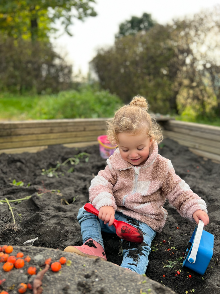 Child in pink jacket plays in a garden, digging soil with a red shovel. Smiling, surrounded by greenery and bushes. Flowers nearby.