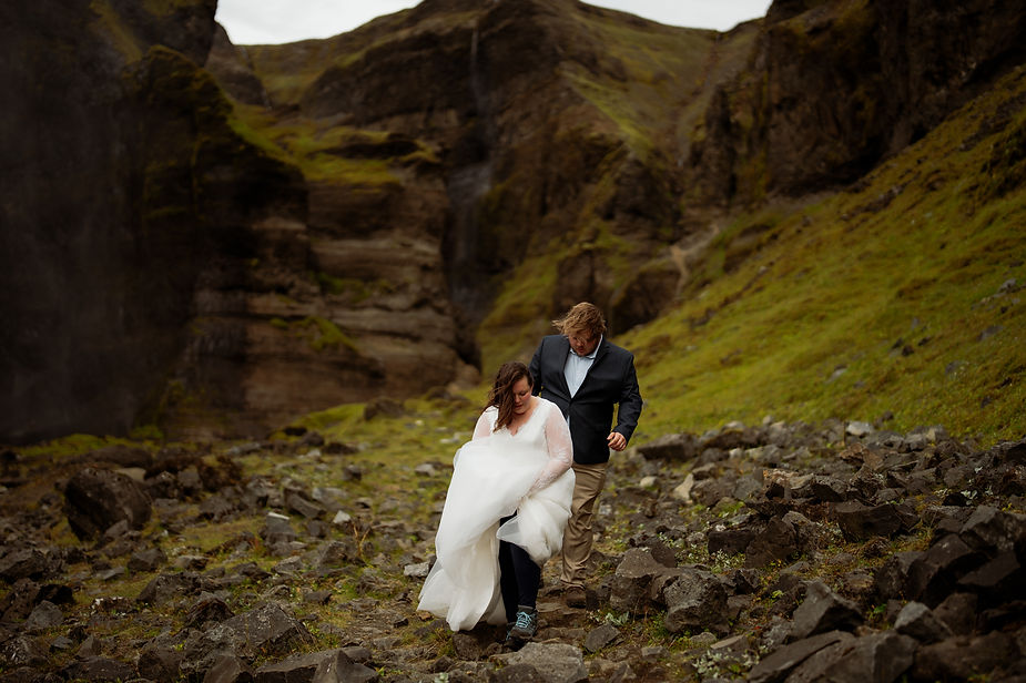 Secret Iceland Elopement Spots: bride and groom walks through rocky, mossy terrain with steep cliffs, showing determination and a sense of adventure.
