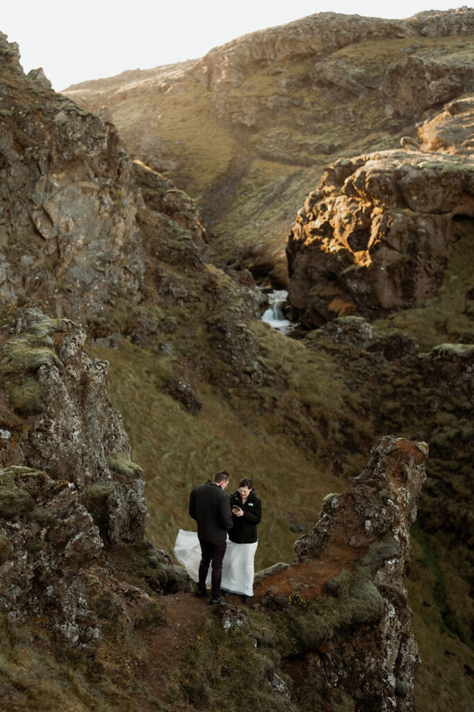 Secret Iceland Elopement Spots: bride and groom stands on a rocky cliffside, surrounded by rugged terrain and soft light, sharing a moment in the wilderness.