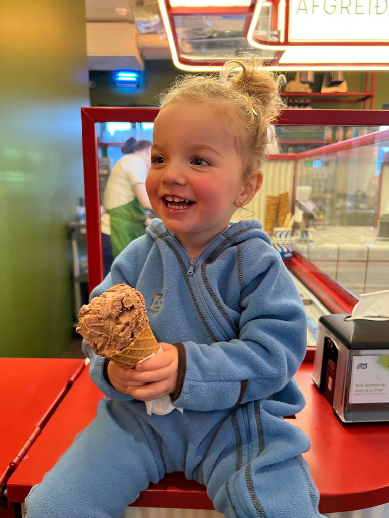 Toddler in blue outfit joyfully holds a chocolate ice cream cone, sitting on a red counter in an iceland shop. Background shows server and equipment.