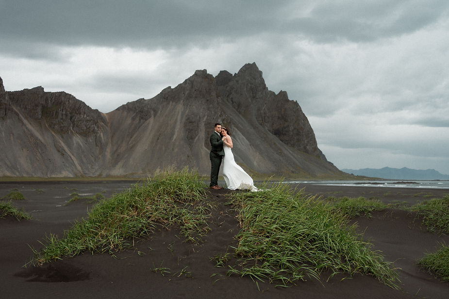 Secret Iceland Elopement Spots: Bride and groom embrace on a grassy mound, set against dramatic gray mountains and sky.