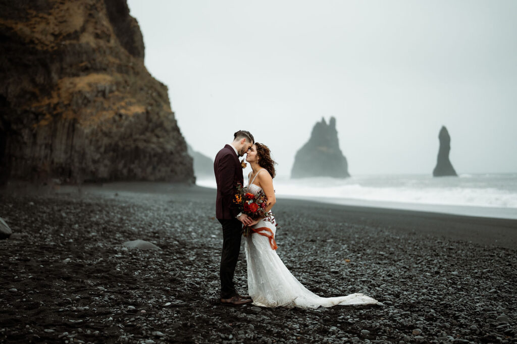 iconic iceland wedding location: Bride and groom embrace on a black sand beach, misty rocks in the background. Bride holds a bouquet.