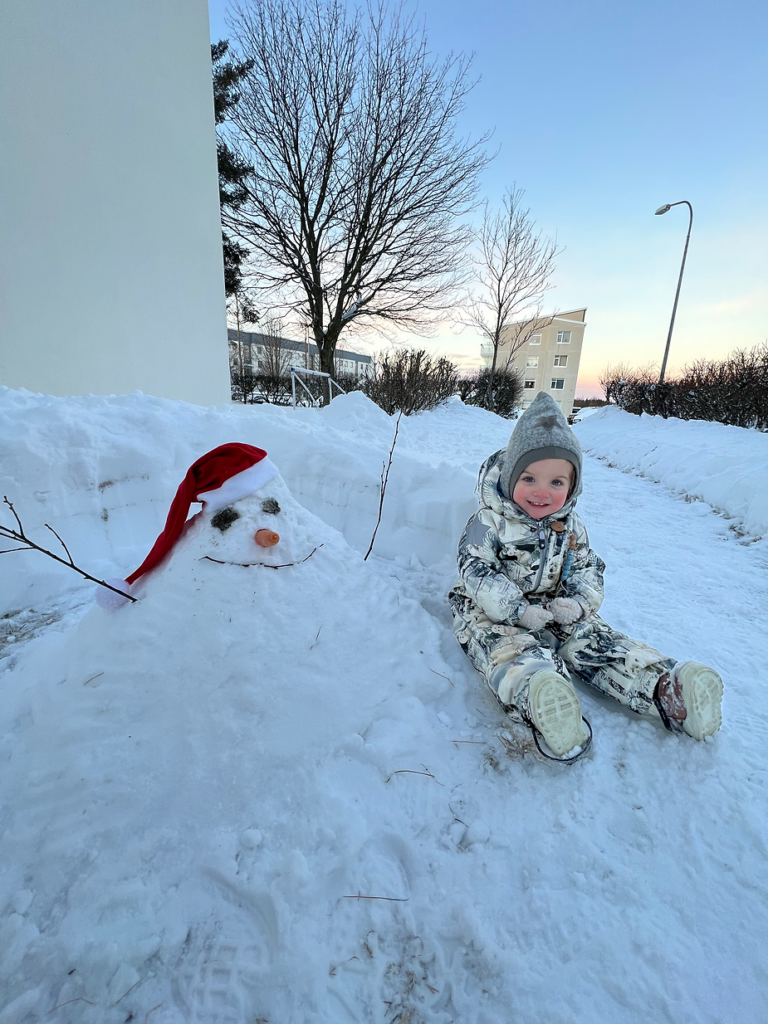Child in winter suit sits on snowy path next to a snowman wearing a red hat. Bare trees and buildings in the sunset background in iceland.