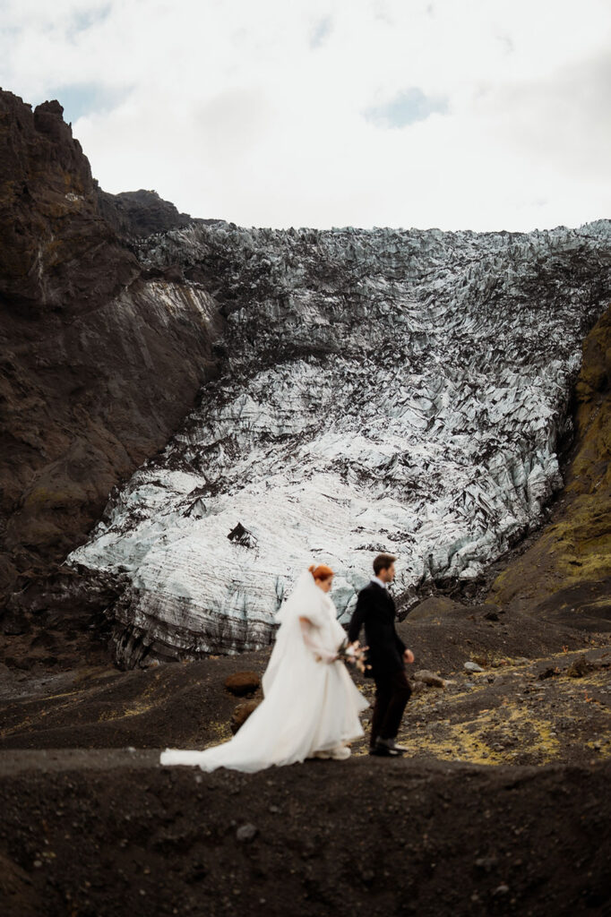 secret iceland elopement spots: Bride and groom walking in front of a dramatic glacier backdrop. The bride wears white and carries flowers. Rugged, earthy landscape.