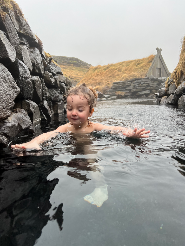 Child enjoying a swim in a natural iceland hot spring, surrounded by stone walls and a grassy, misty landscape. Joyful mood.