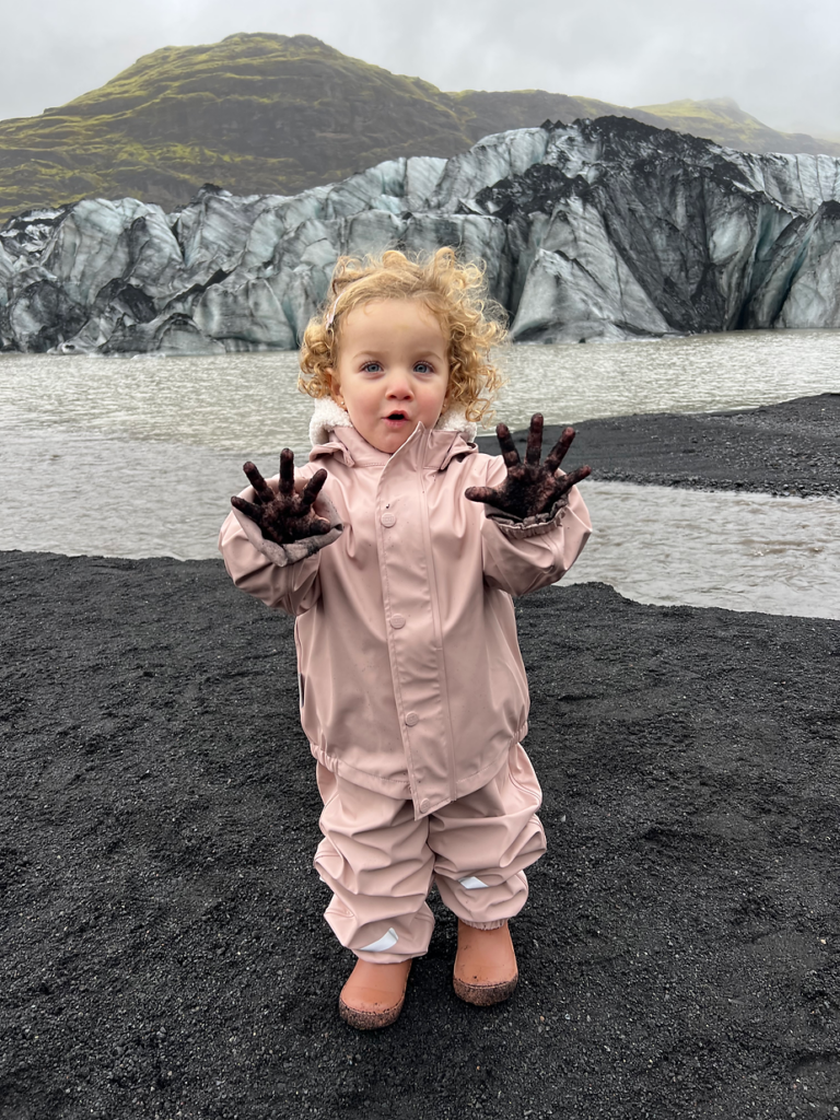 Toddler in pink raincoat plays with muddy hands by an iceland glacial lagoon, with misty mountains and ice in the background. Moody, overcast day.