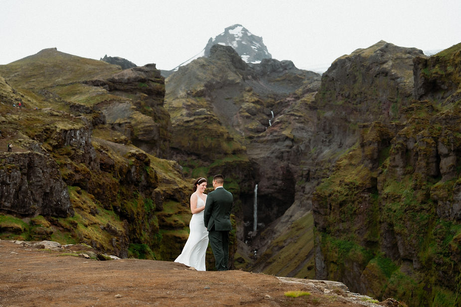 A couple in formal attire stands on a rocky cliff with a waterfall and mountains in the background, creating a serene and romantic scene.