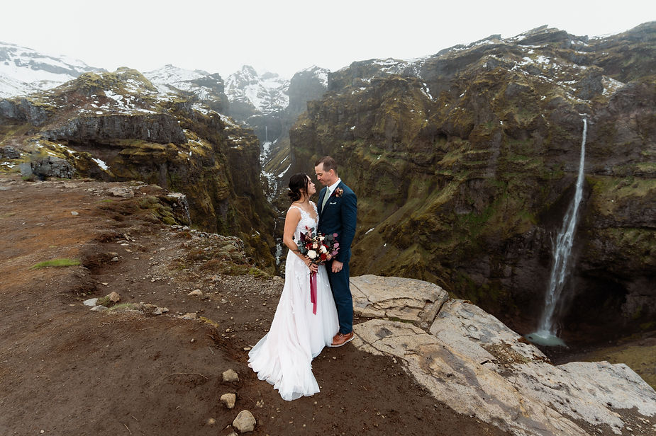 secret iceland elopement spots: Bride in a white gown and groom in a blue suit stand on a rocky cliff with a waterfall and snow-covered mountains in the background.